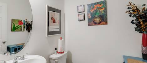 Une salle de bain avec un lavabo, des toilettes et un miroir, tous de couleur blanche. Des tableaux encadrés ornent le mur, et un vase rempli de fleurs y est également posé.