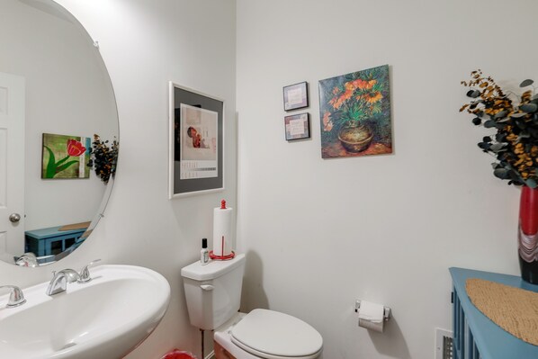 Une salle de bain avec un lavabo, des toilettes et un miroir, tous de couleur blanche. Des tableaux encadrés ornent le mur, et un vase rempli de fleurs y est également posé.