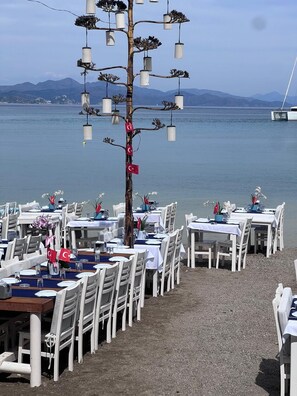 A seaside restaurant with tables and chairs arranged for dining, overlooking a calm body of water and distant mountains.