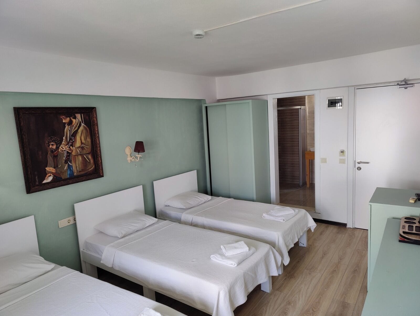Une chambre d’hôtel avec trois lits simples, une armoire, un tableau au mur et une porte donnant sur une salle de bain.