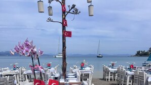 A seaside restaurant with tables set for dining, featuring Turkish flags and orchid decorations.