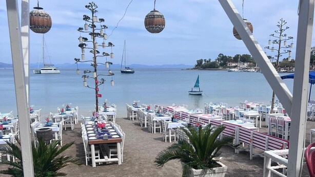 A seaside terrace with tables and chairs, a view of the sea, and hanging baskets.