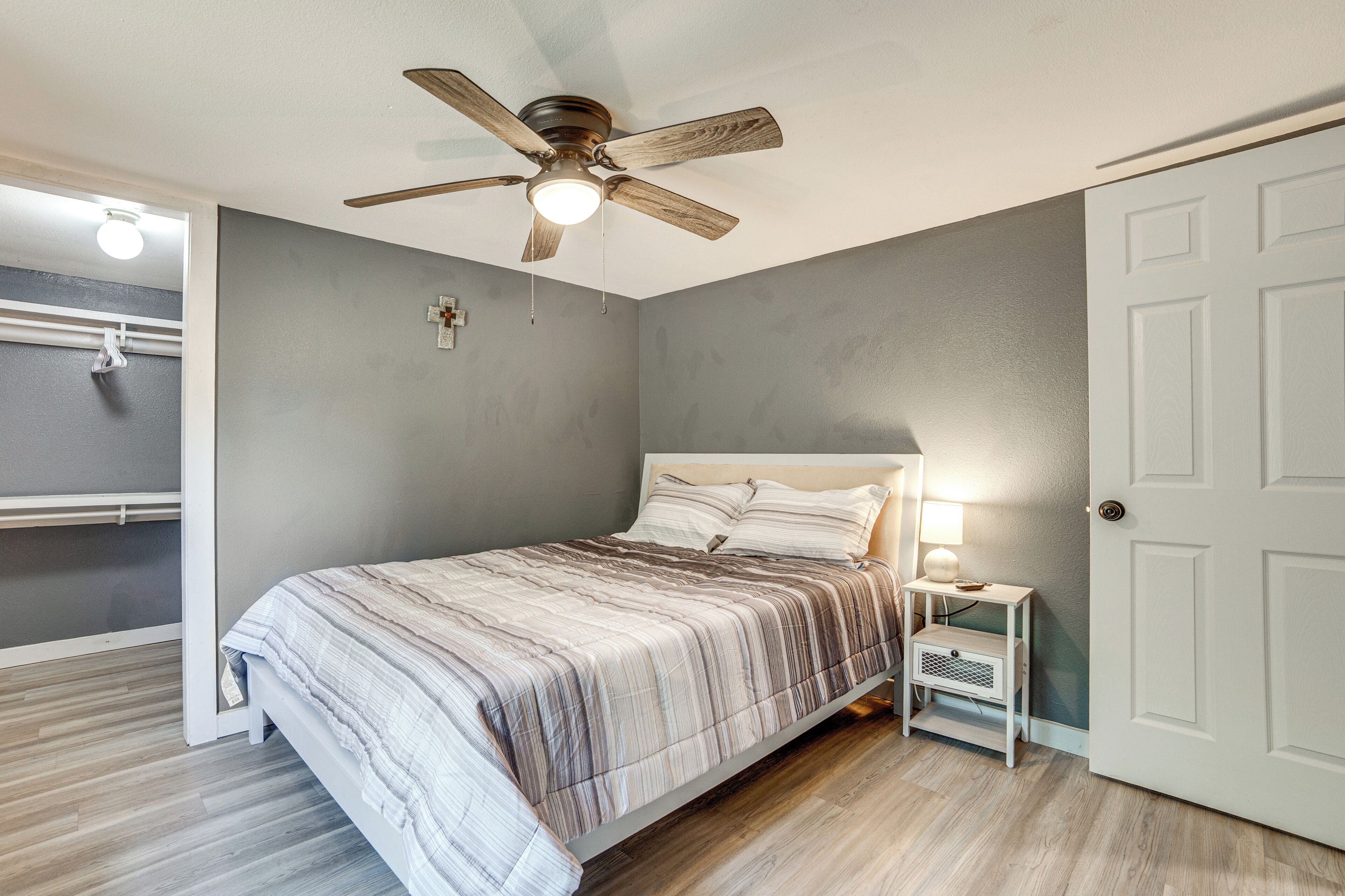 Un dormitorio con cama, ventilador de techo, mesita de noche con lámpara y una puerta.
