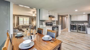 A modern kitchen with white cabinets, a dining area with a wooden table set for four, and a living room with a sofa and coffee table.