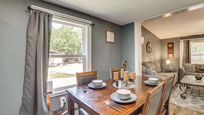 A dining area with a wooden table set for four, a sofa, and a window with curtains. - Lake Tawakoni Basecamp: Covered Patio + Fire Pit! (Wills Point)