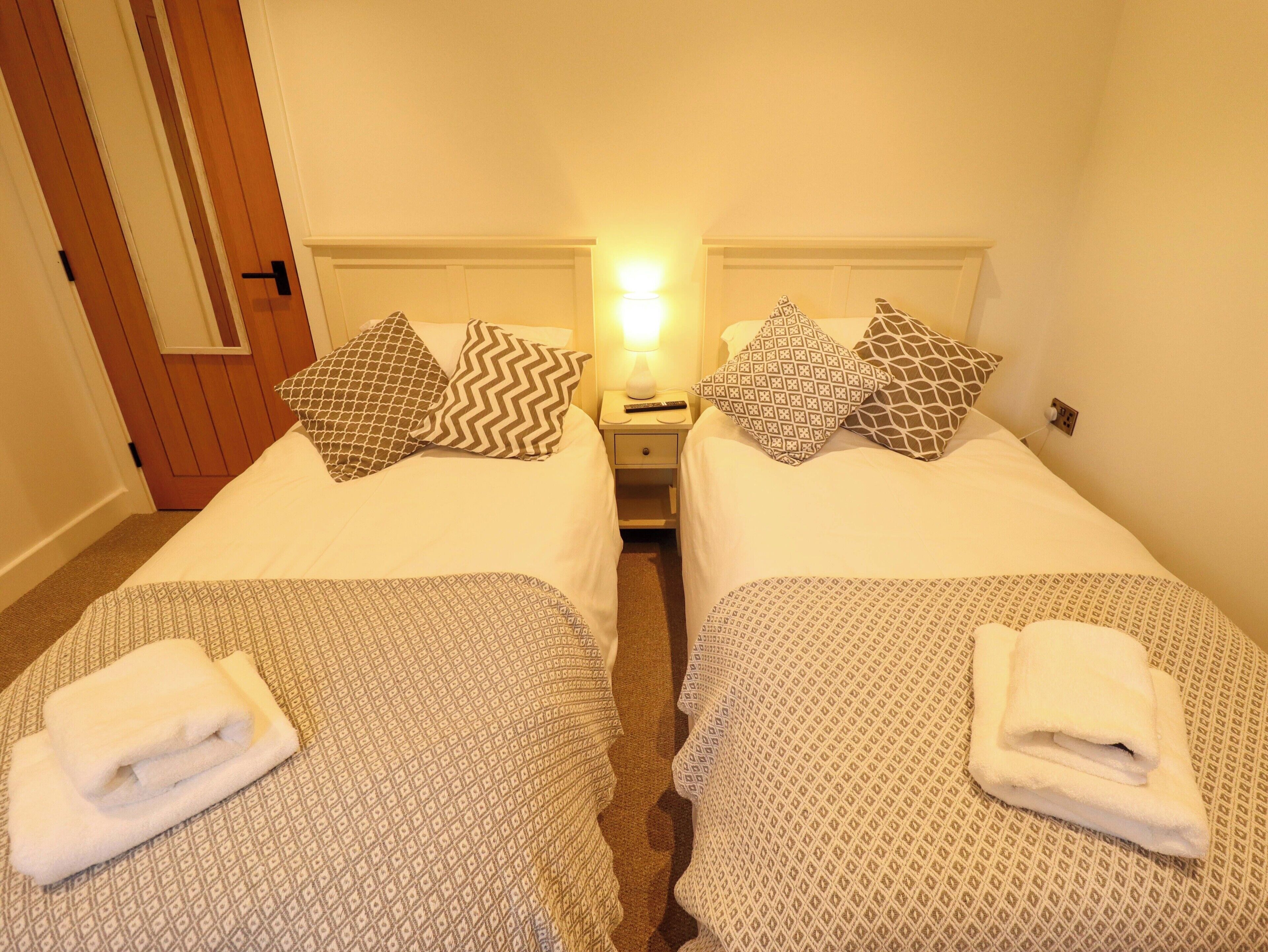 A hotel room with two beds, each with patterned bedding and pillows, a nightstand with a lamp, and a door with a wooden panel.