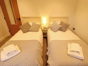 A hotel room with two beds, each with patterned bedding and pillows, a nightstand with a lamp, and a door with a wooden panel. - The Hen House (King's Lynn)