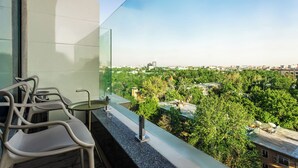 Un balcón con barandilla de vidrio, una mesa y dos sillas, con vistas a un paisaje urbano de edificios y árboles.