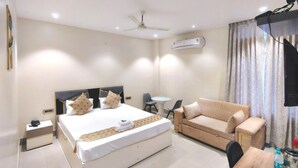 A hotel room with a bed, a sofa, a small table, a ceiling fan, an air conditioning unit, and a television. - Hotel H - 22 (Noida)