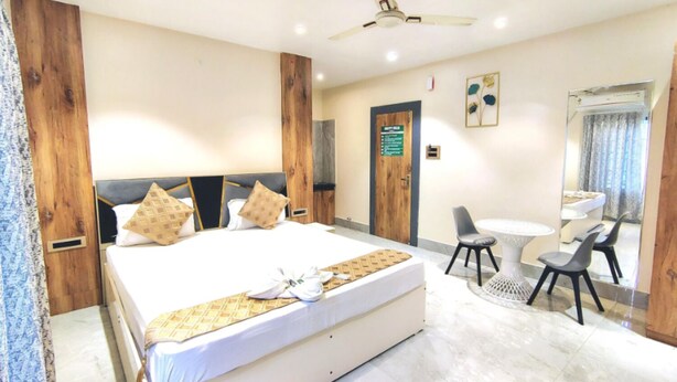 A hotel room with a large bed, a desk, a chair, a ceiling fan, and a bathroom visible through a glass door. - Hotel H - 22 (Noida)