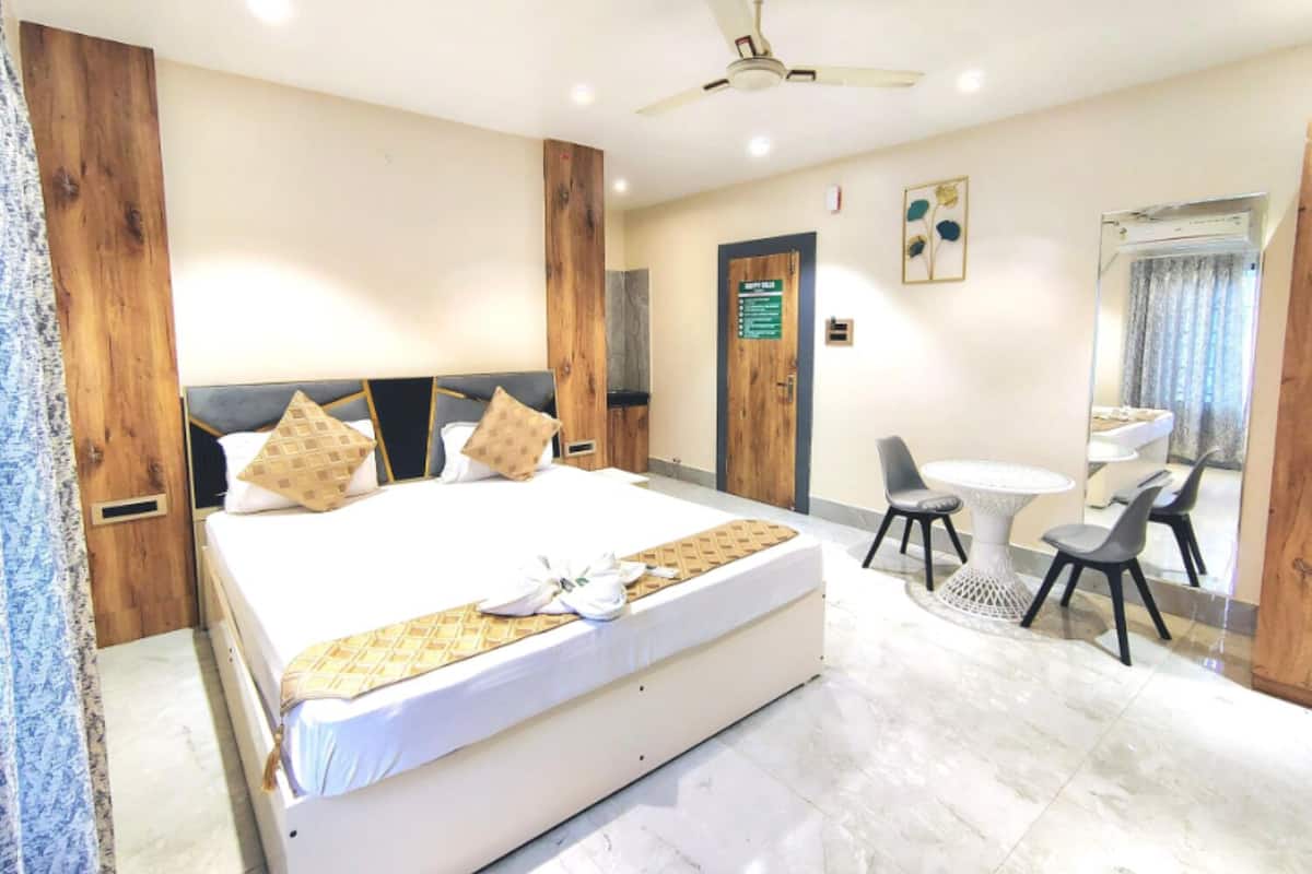 Habitación de hotel con una cama grande, un escritorio, una silla, un ventilador de techo y un baño visible a través de una puerta de vidrio.