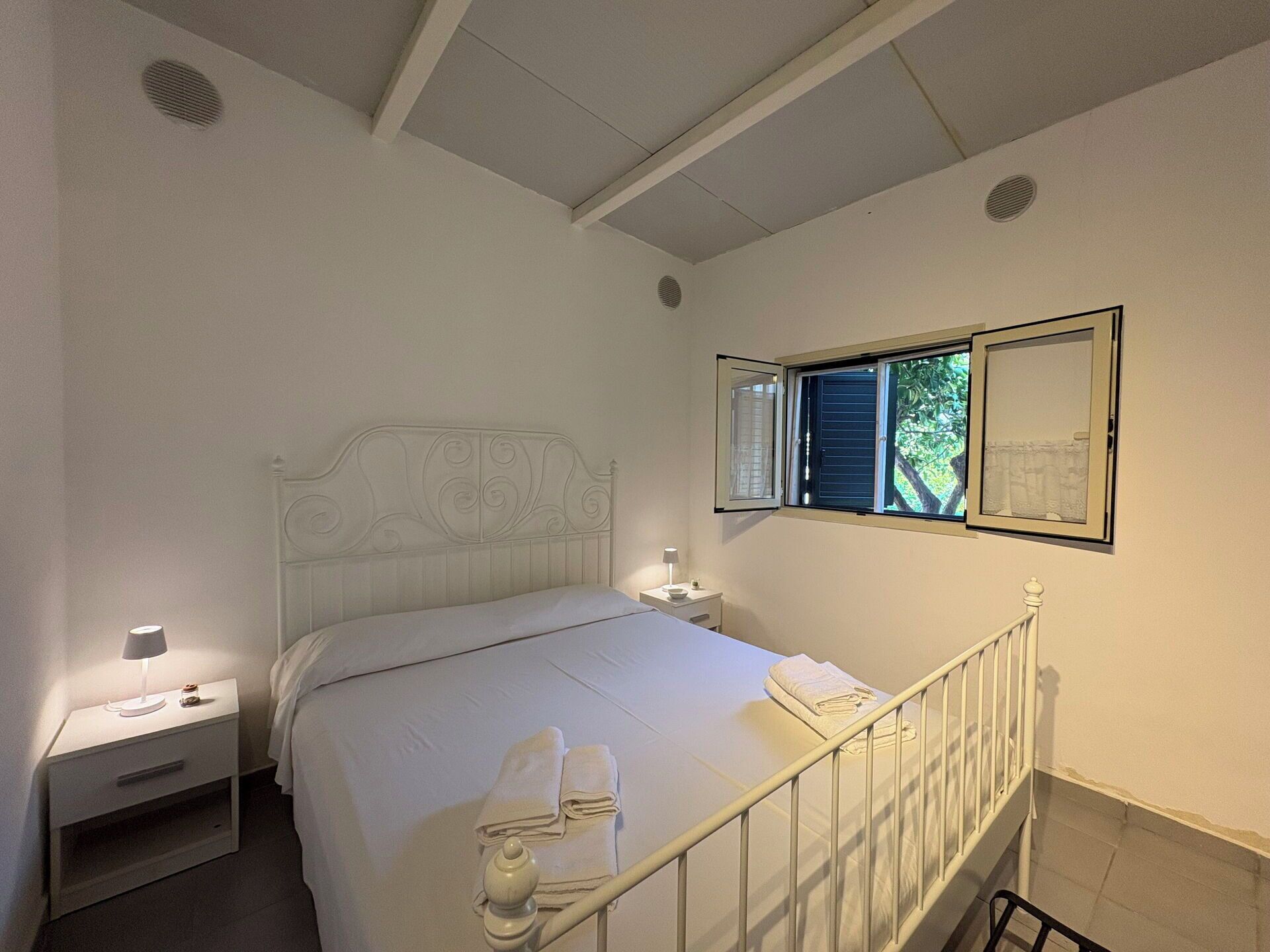 A bedroom with a bed, two bedside tables, a window with blinds, and a wall-mounted picture.