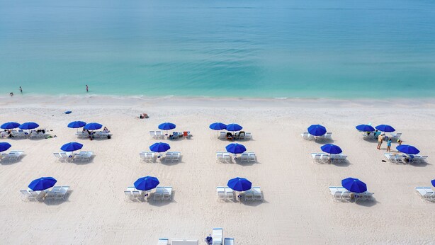 A beach with rows of white lounge chairs and blue umbrellas, some occupied, under a clear sky. - West Bay Club first floor Coach Home with Lake View (Estero)