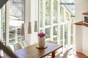 A dining area with a wooden table, white chairs, a vase with flowers, and large windows with a view of greenery.