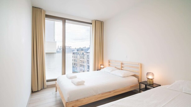 A hotel room with a large window, a bed, a bedside table with a lamp, and a second bed. - City Rhythm Abode for 10 in Antwerp (Antwerp)