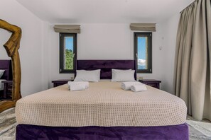Ein großes Bett mit violettem Kopfteil, zwei Nachttische und Blick ins Freie durch Fenster.