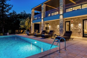 A modern outdoor pool area with lounge chairs, a clear pool, and a stone building with a balcony. - BRAND NEW LUXURY 4-BEDROOM VILLA IN CENTRAL LOCATION -70 METERS FROM THE SEA! (Nydri)