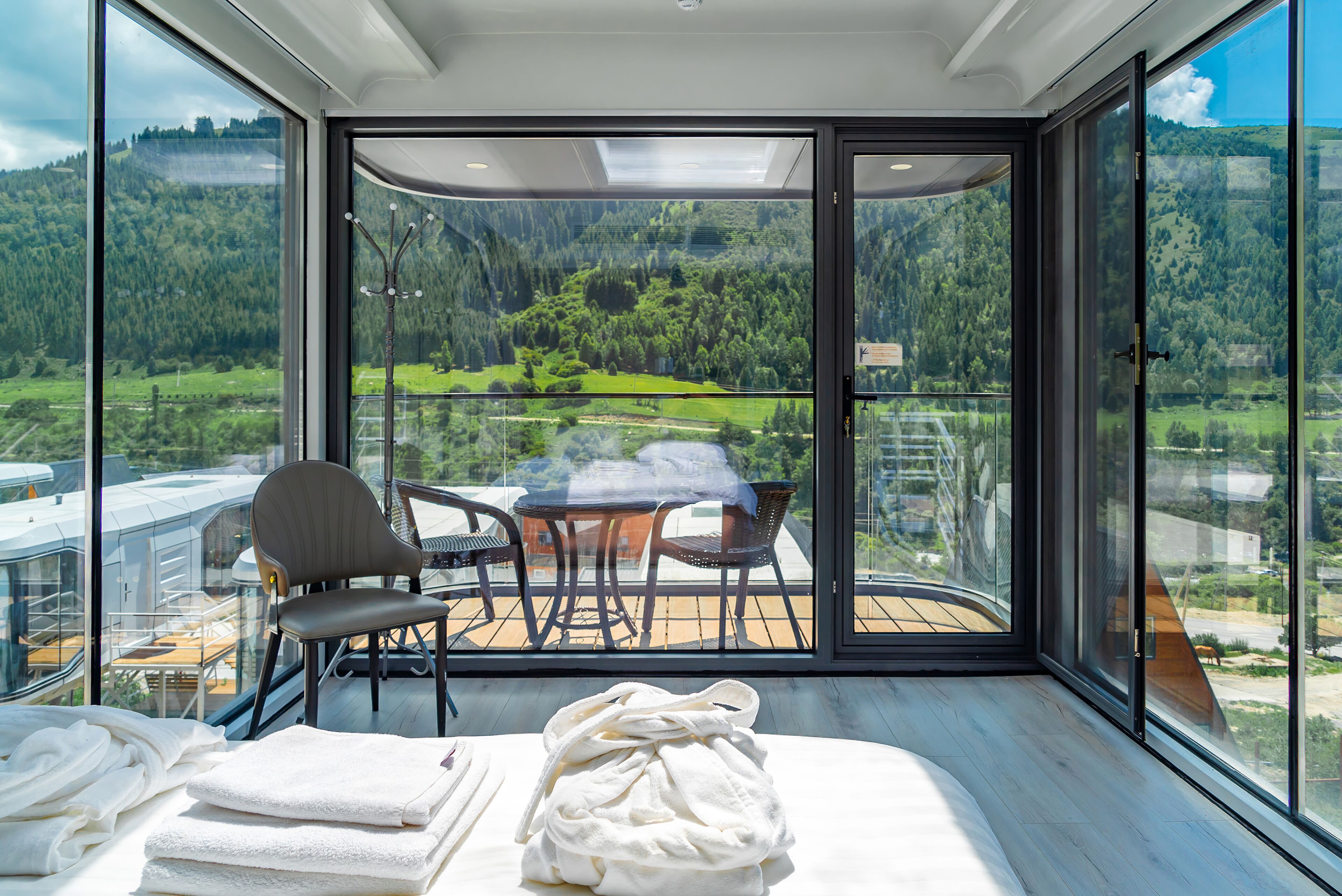 A balcony with a table and chairs, a view of a forested landscape, and a pile of towels.