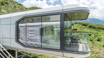 Una cabaña moderna, con paredes de vidrio y una escalera que conduce a un balcón, con vistas a un paisaje montañoso.