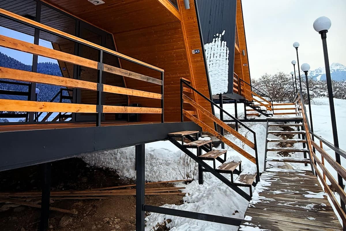 A modern wooden cabin with a sloped roof, a wooden deck, and a view of snow-covered mountains.