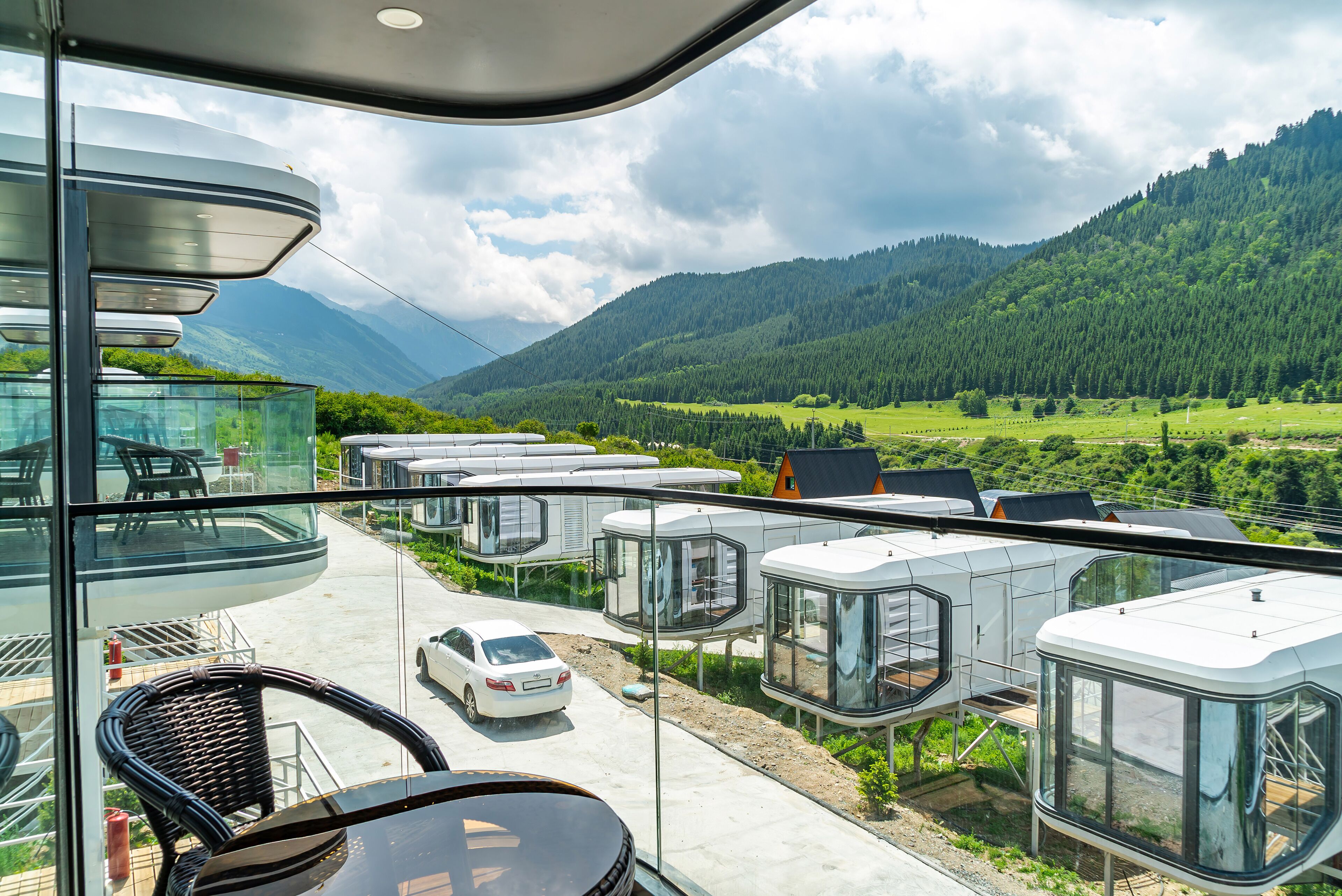 A modern building with a glass exterior, a parking area with a white car, and a view of green hills and mountains.