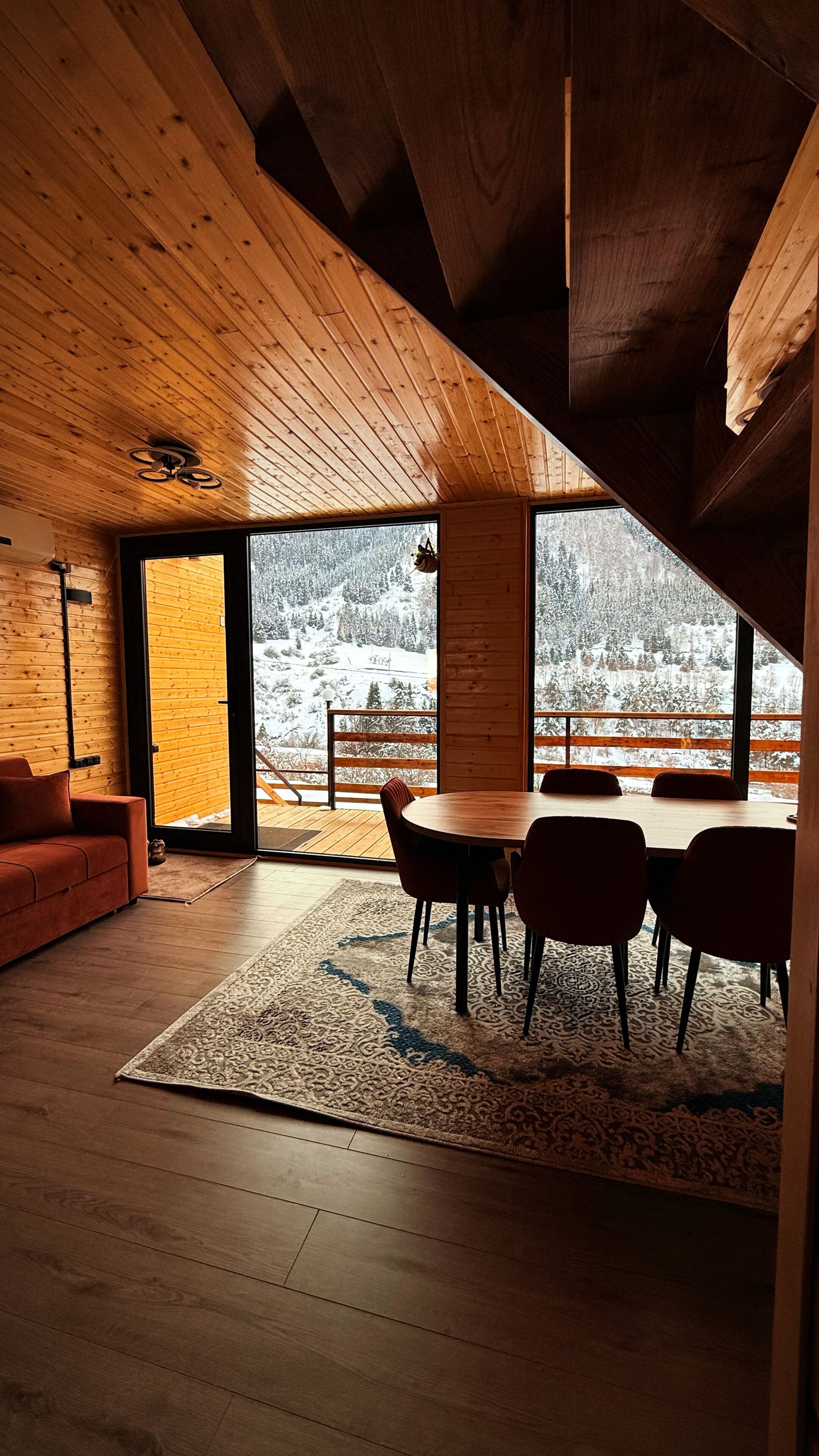 Un interior moderno con techo de madera, una zona de comedor con sillas, un sofá y vistas a un paisaje nevado a través de amplios ventanales.