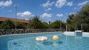 Une piscine aux eaux cristallines, agrémentée de deux bouées gonflables et d’un ballon de plage, le tout entouré d’un mur de pierre et d’arbres.