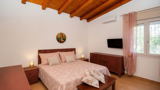 Une chambre avec un plafond en bois, un lit recouvert d’une couvre-lit à motifs floraux, une table de chevet avec une lampe, une commode et un téléviseur fixé au mur.