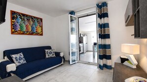 A living room with a blue sofa, a glass door leading to a kitchen, and a colorful painting on the wall. - Stunning  villa for 4 guests with A/C, WIFI, private pool, TV, patio and panoramic view (Marina Di Ragusa)