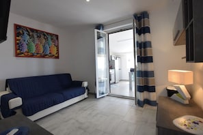 Ein Wohnzimmer mit einem blauen Sofa, einem Gemälde, einer Glastür, die in eine Küche führt, und einer Lampe.