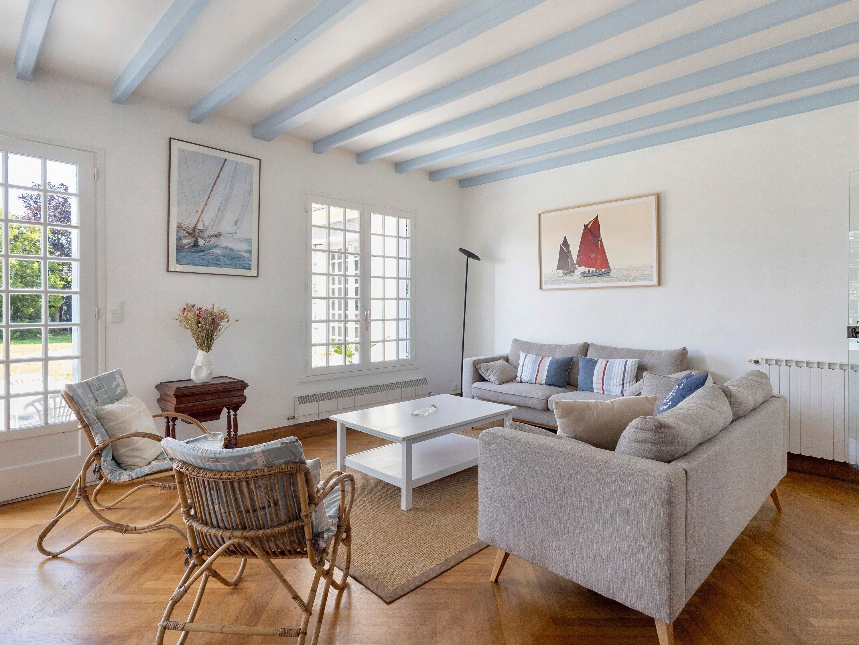 Ein Wohnzimmer mit einer Couch, einem Sessel, einem Couchtisch und einer Stehlampe. Es gibt Fenster mit weißen Rahmen und ein Segelboots-Gemälde an der Wand.