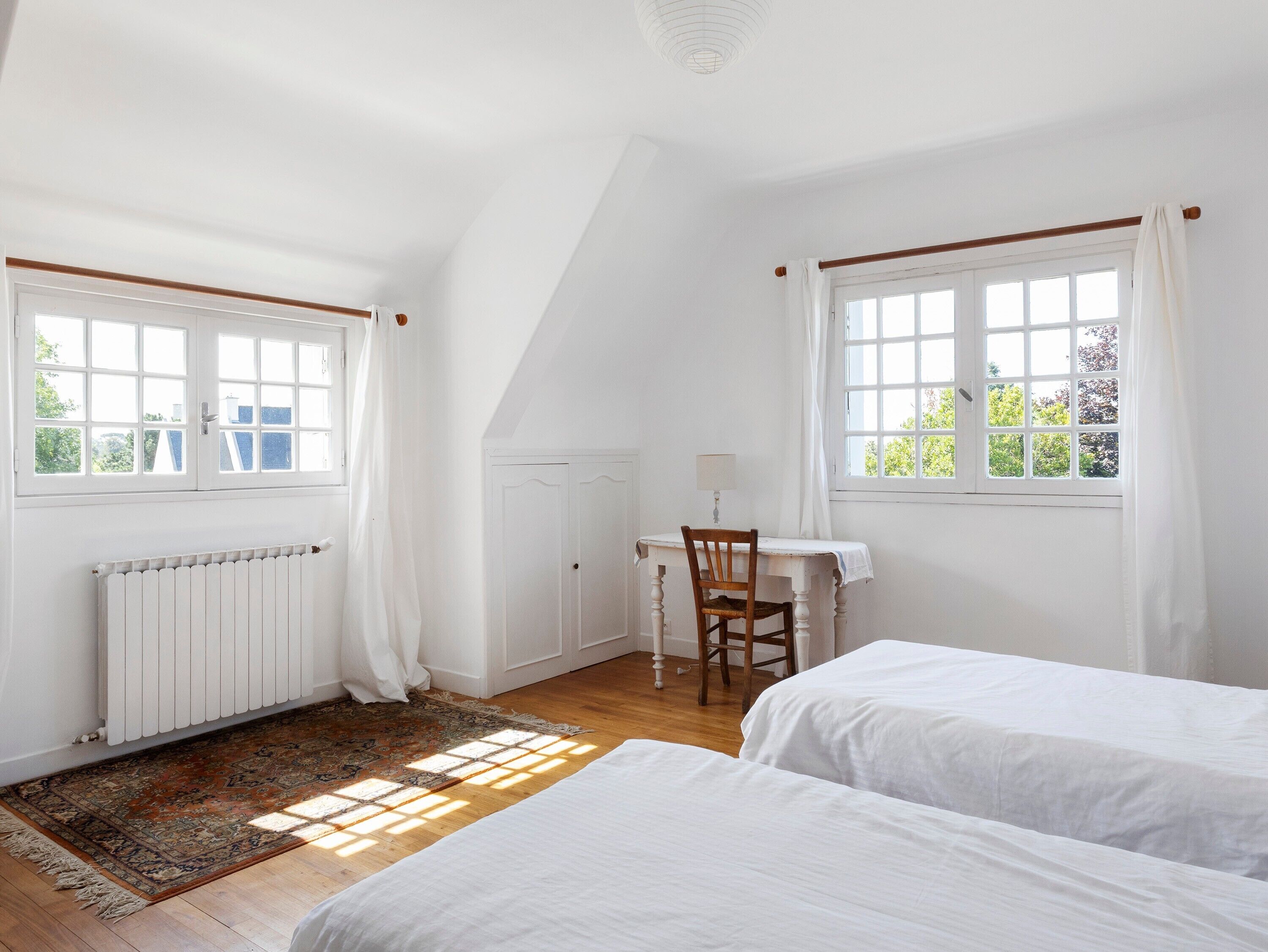 Une chambre avec deux lits, un bureau avec une chaise, un radiateur et une fenêtre avec des rideaux.