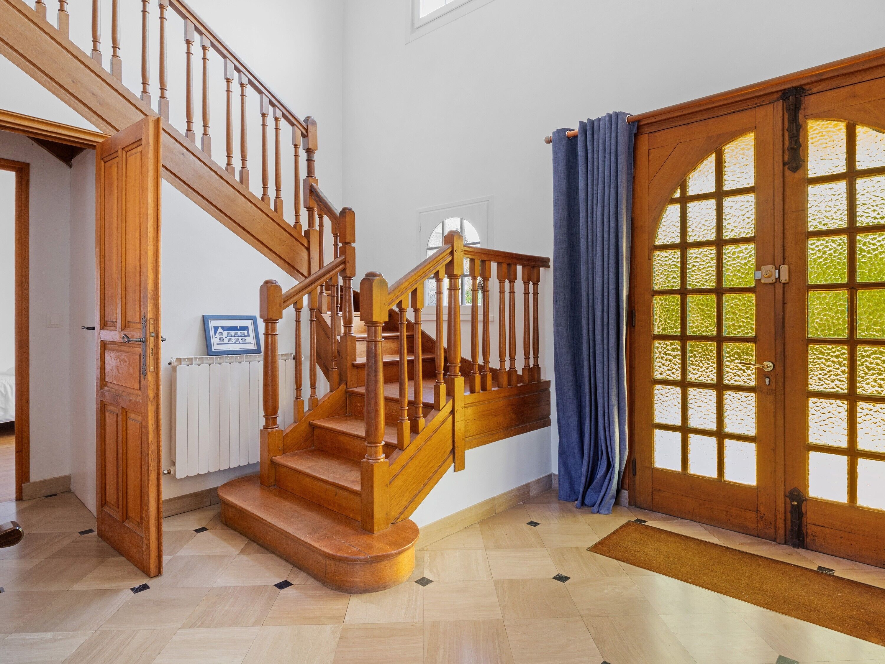 Un escalier en bois menant à l’étage supérieur, une porte avec un panneau de verre et un radiateur fixé au mur.