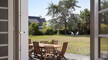 An open door leading to an outdoor patio with a wooden table and chairs, surrounded by a stone floor, with a view of a house and trees in the background.