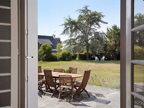 An open door leading to an outdoor patio with a wooden table and chairs, surrounded by a stone floor, with a view of a house and trees in the background.