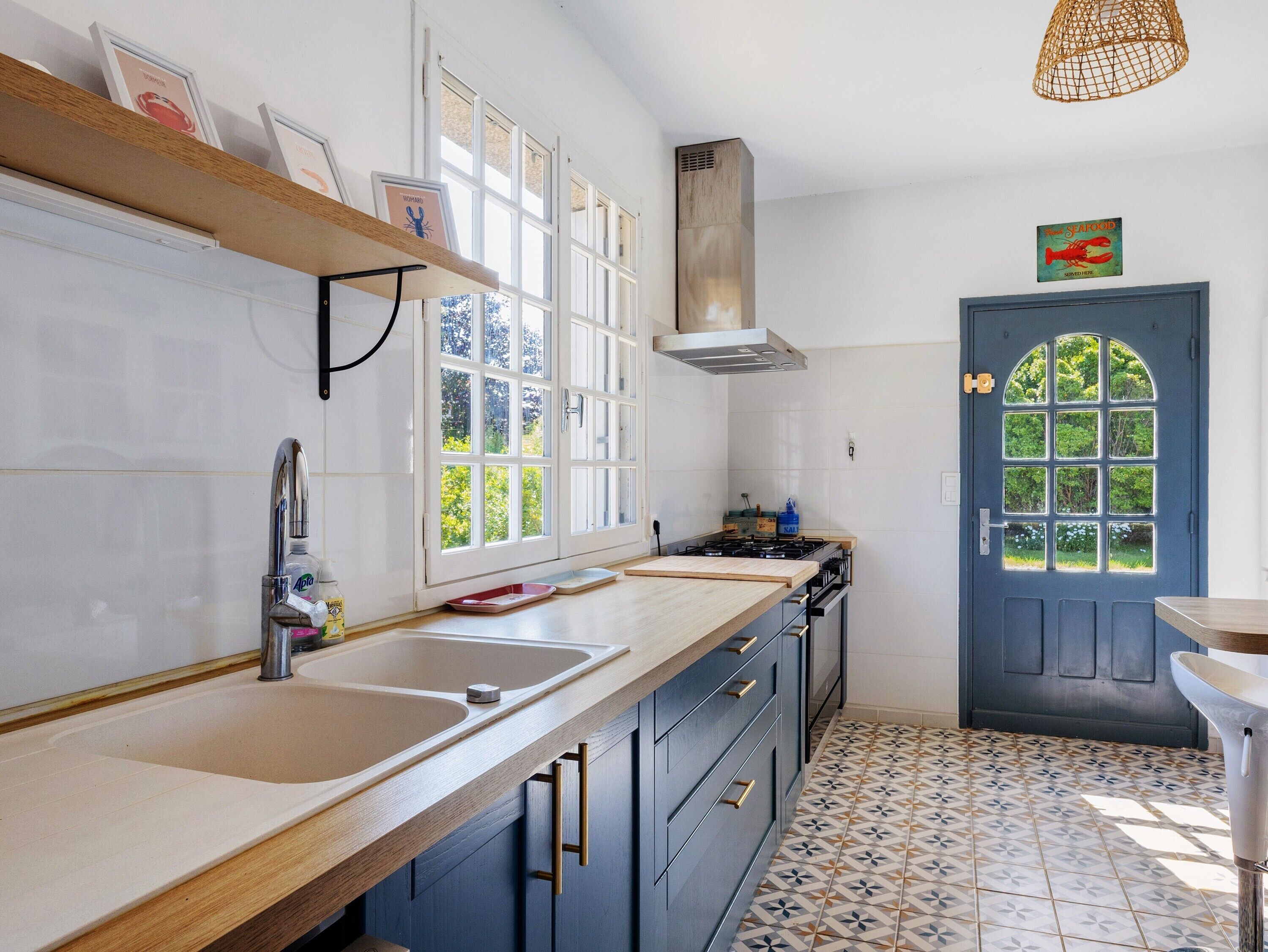 A kitchen with a double sink, wooden countertop, and a stove. There is a blue door with a window and a shelf with framed pictures.