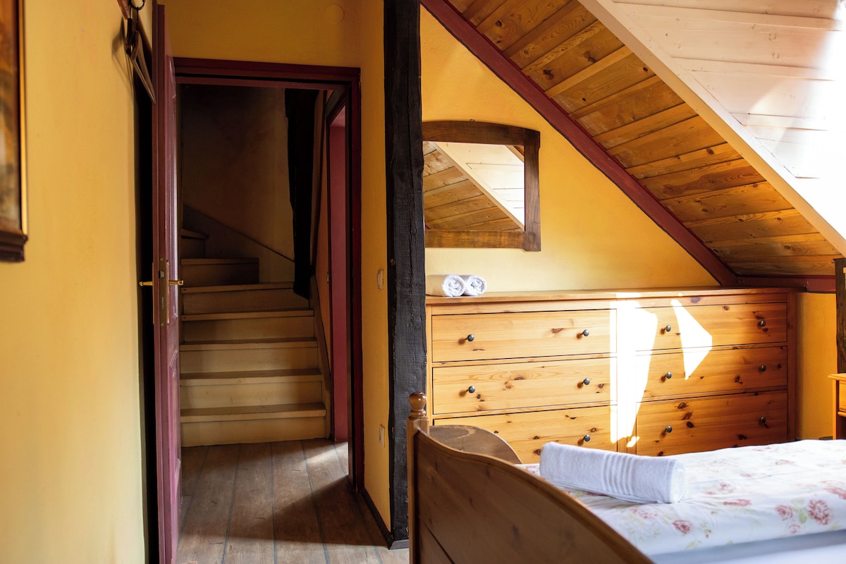 Une chambre à coucher avec une tête de lit en bois, un lit recouvert d’une couvre-lit à motifs floraux, une commode en bois et un escalier menant à un étage supérieur.