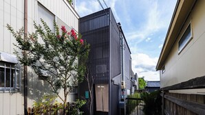 En smal bygningsgang med en moderne bygning med lodrette lister, et træ med lyserøde blomster og en traditionel japansk port.