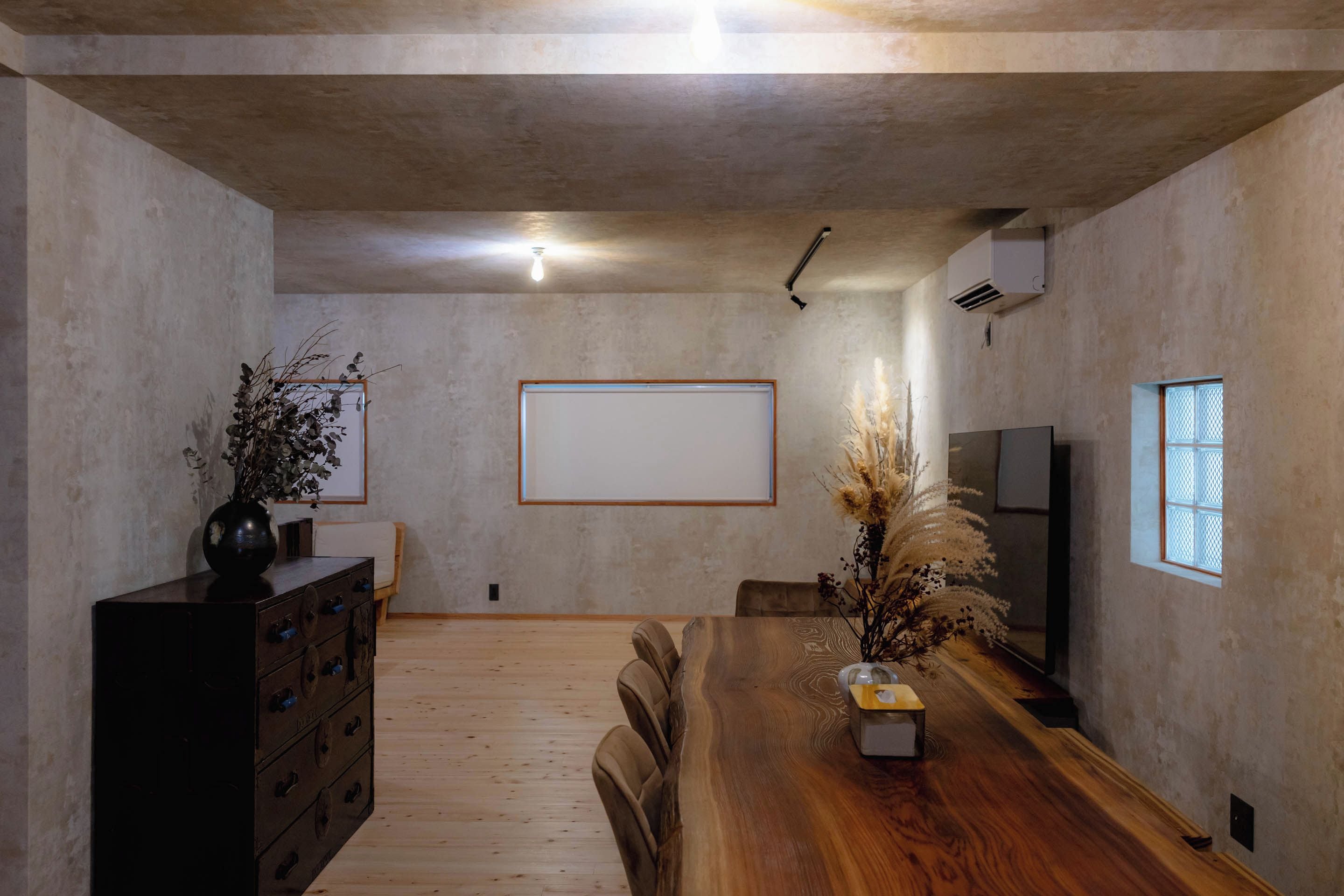 A minimalist room with a wooden dining table, a dark chest of drawers, a whiteboard, and a window with blinds.