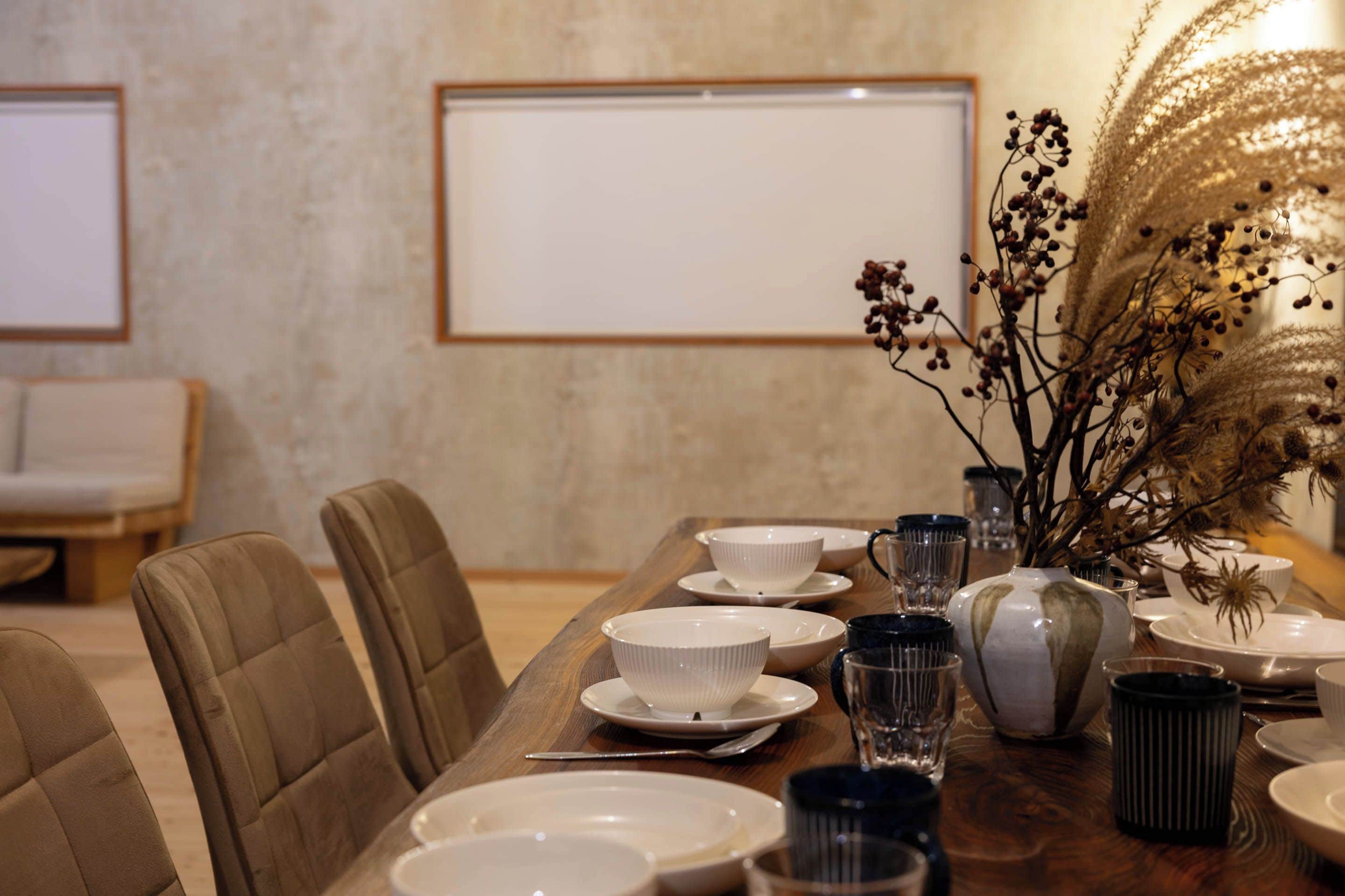 Una mesa de comedor dispuesta con platos, cuencos y vasos, junto a sillas y un florero con ramas secas.
