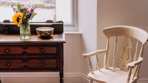 Un comò in legno con cassetti, una sedia bianca, un vaso con fiori e una ciotola decorata su un davanzale.