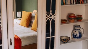 Una stanza con un letto, cuscini, una libreria piena di libri e oggetti decorativi, e una finestra con un pannello decorativo.