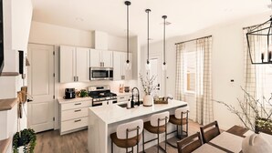 A modern kitchen with white cabinetry, a central island, bar stools, pendant lighting, and a dining area with a table and chairs.