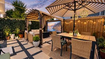 An outdoor patio with a pergola, seating, a table with candles, and a TV screen displaying a scenic view.