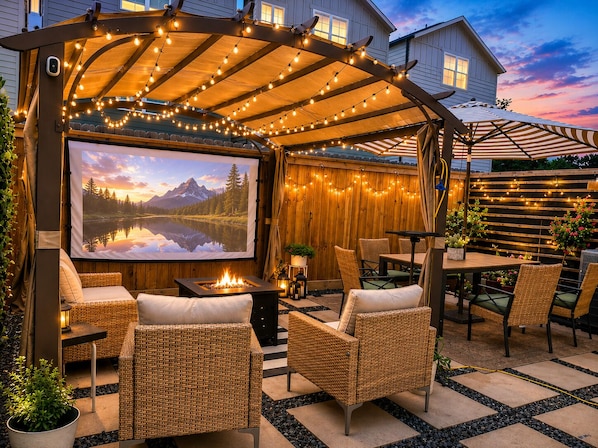 Eine Außenpatio mit Korbmöbeln, einer Feuerstelle, Lichterketten und einem großen Bildschirm, der eine schöne Landschaft zeigt.