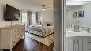 Ein Schlafzimmer mit Bett, Kommode, Fernseher und einem Badezimmer mit Waschbecken und Spiegel.