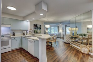 A modern kitchen with wooden flooring, white cabinetry, and a dining area with wicker furniture. - Sunset Rentals | Colonnade Club 147 (Hilton Head Island)