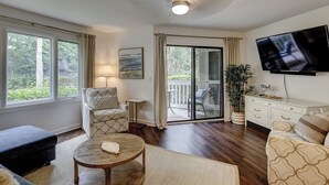 Ein Wohnzimmer mit einer Couch, einem Sessel, einem Couchtisch, einem Fernseher und einer Schiebetür mit Glas, die auf einen Balkon führt.