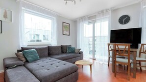 Una sala moderna con un sofá seccional, una pequeña mesa de centro de madera, un comedor con sillas, un televisor de pantalla plana y un ventanal con cortinas.