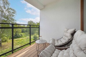 Un balcone con vista sul verde, una sedia in vimini, un tavolino rotondo con una caraffa di vetro e un divano bianco imbottito.
